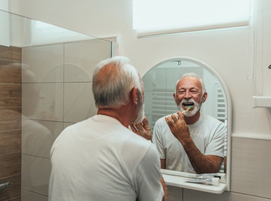 older man brushing his teeth in front of a bathroom mirror