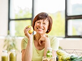 An older woman seated at a table and holding a whole apple while smiling to show off her new dental implants