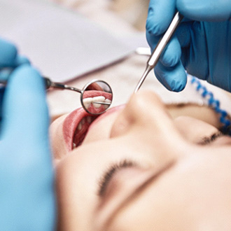 Blue-gloved hands holding dental instruments examining woman’s teeth