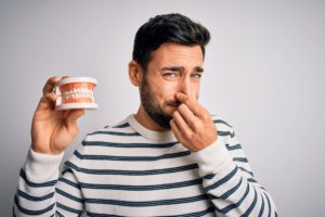 Man in striped sweater holding his nose closed with one hand and a set of dentures in the other